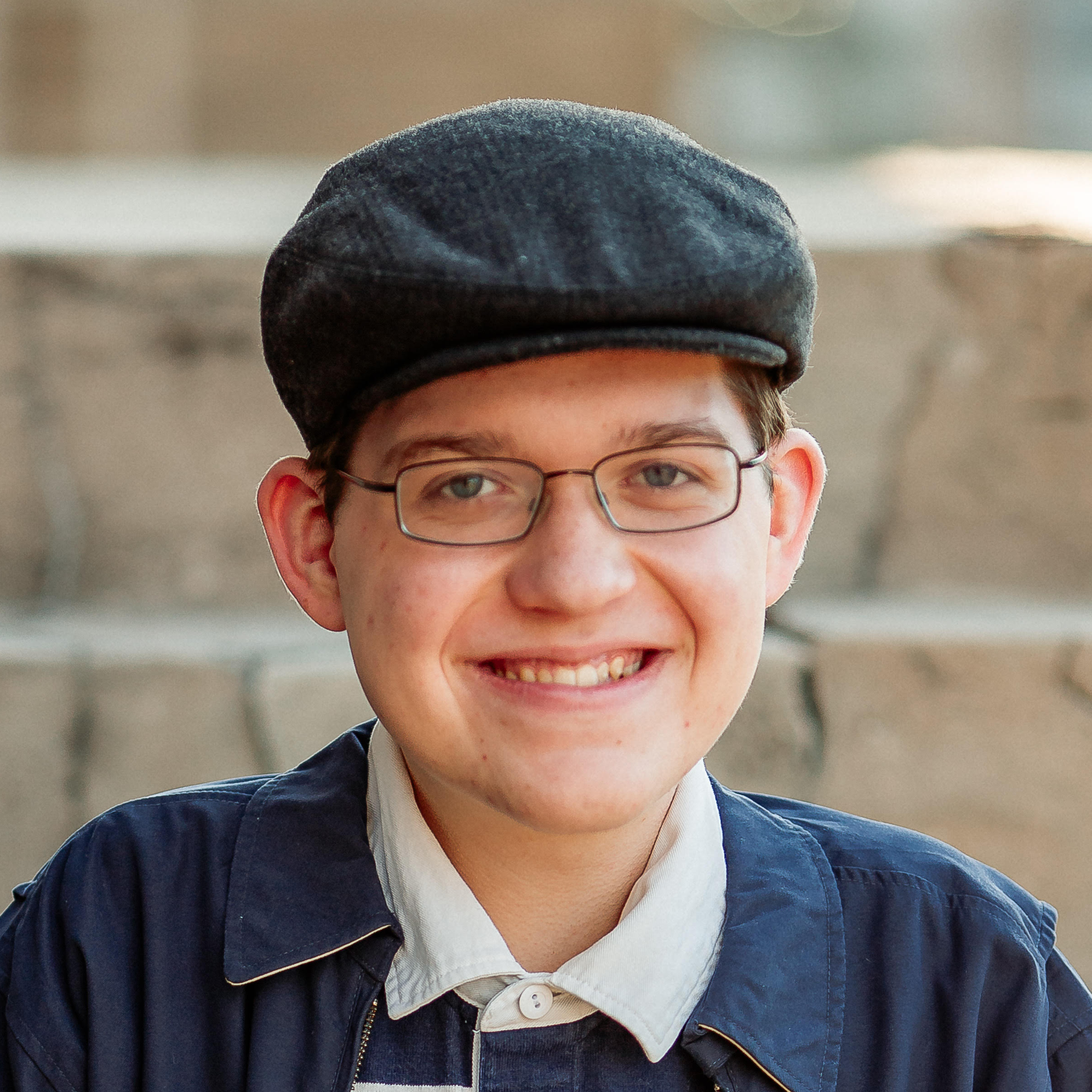 A person with glasses, a hat, and a blue jacket smiles at the camera while sitting outdoors.
