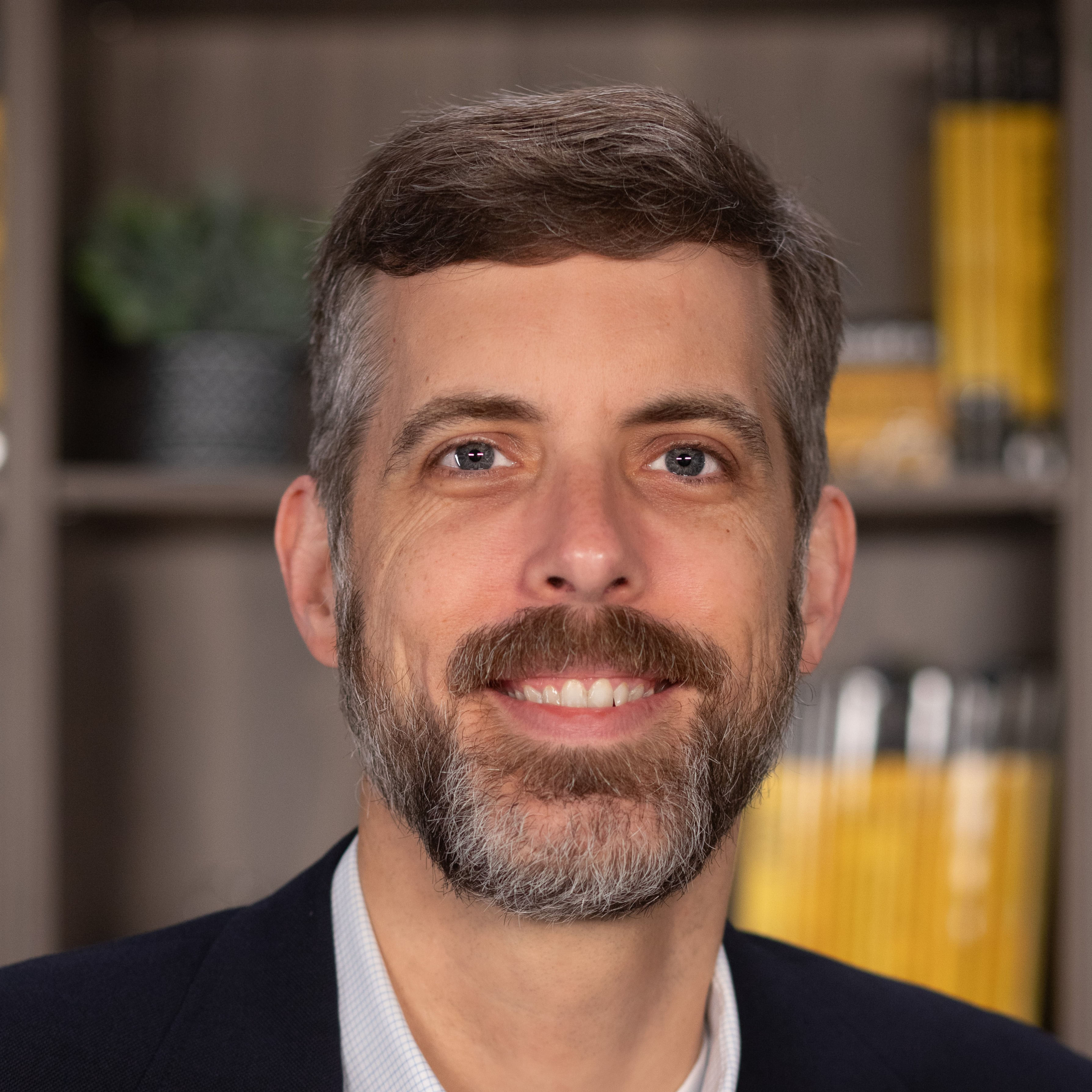 A man with short brown hair and a beard smiles, standing in front of shelves filled with yellow and black books.