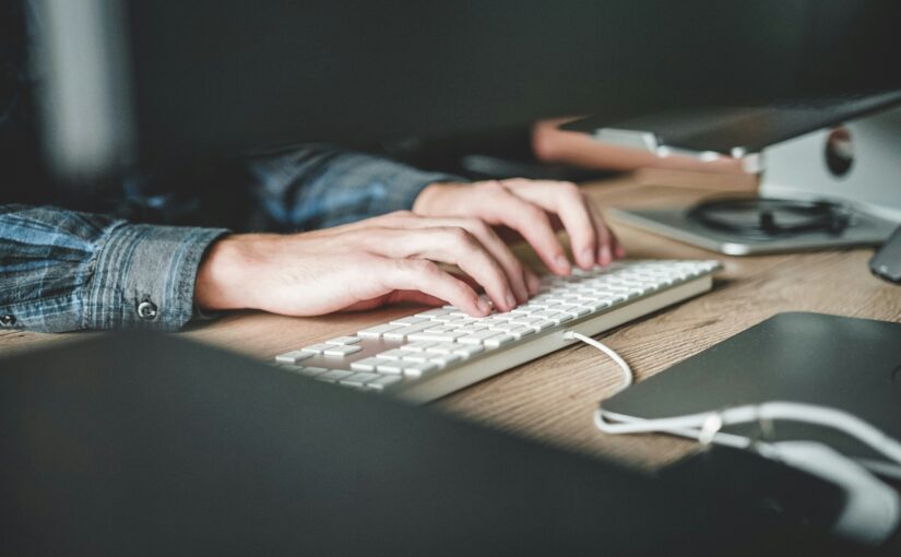 A person wearing a denim shirt is typing on a white keyboard at a wooden desk with a computer and other electronic devices.